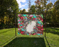 A large green, teal and pink beaded tapestry depicting a pixelated  supernovae and gas rings made by the Hubble Space Telescope. The beaded tapestry is hung from a metal frame on a grassy lawn.
