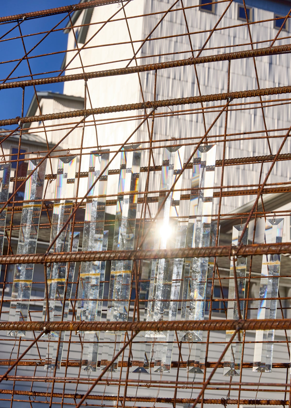 A grid of rusty steel rebar holds rows of clear, vertically hanging rectangular crystal prisms that catch and refract bright sunlight against a soft-focus background of a white industrial building.
