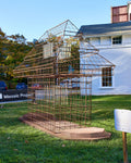 A large outdoor sculpture sits on a grass lawn. The artwork is a life-sized, open outline of a Fotomat--a pagoda-roofed photo kiosk constructed from a grid of rusty steel rebar and mesh, set in front of a white wooden building at the Wassaic Project.