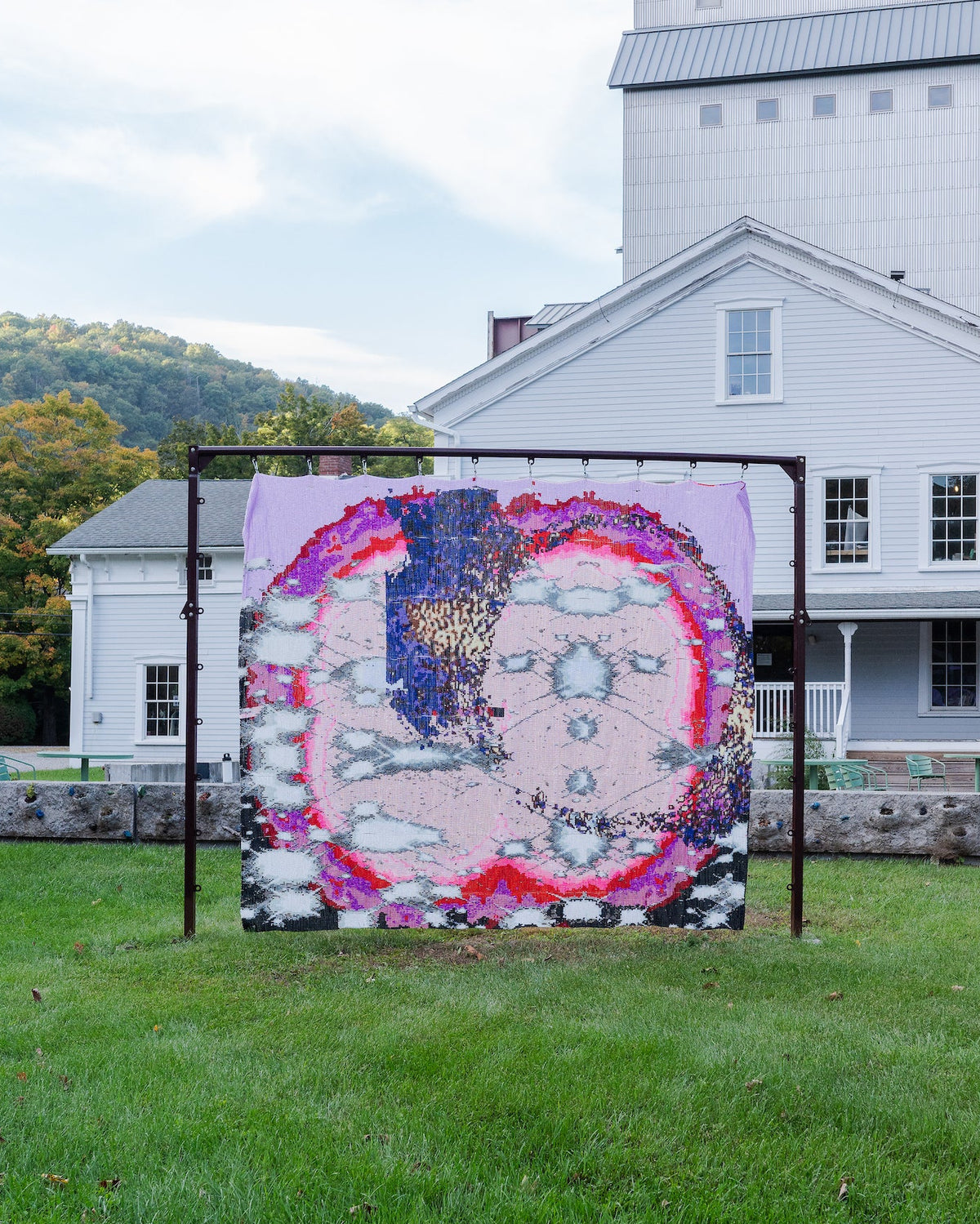 A large pink and multicolor beaded tapestry depicting a pixelated  supernovae and gas rings made by the Hubble Space Telescope. The beaded tapestry is hung from a metal frame on a grassy lawn in front of a white historic building and a grain elevator.